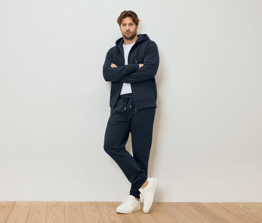 Un homme aux bras croisés porte un sweat à capuche zippé bleu marine, un pantalon de survêtement bleu marine et des baskets blanches, debout devant un mur blanc.