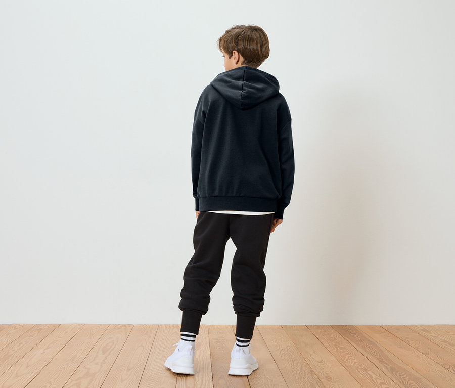 Un enfant aux cheveux bruns se tient dos à la caméra, portant un sweat à capuche noir, un pantalon de jogging noir, des chaussettes blanches à rayures et des chaussures blanches.