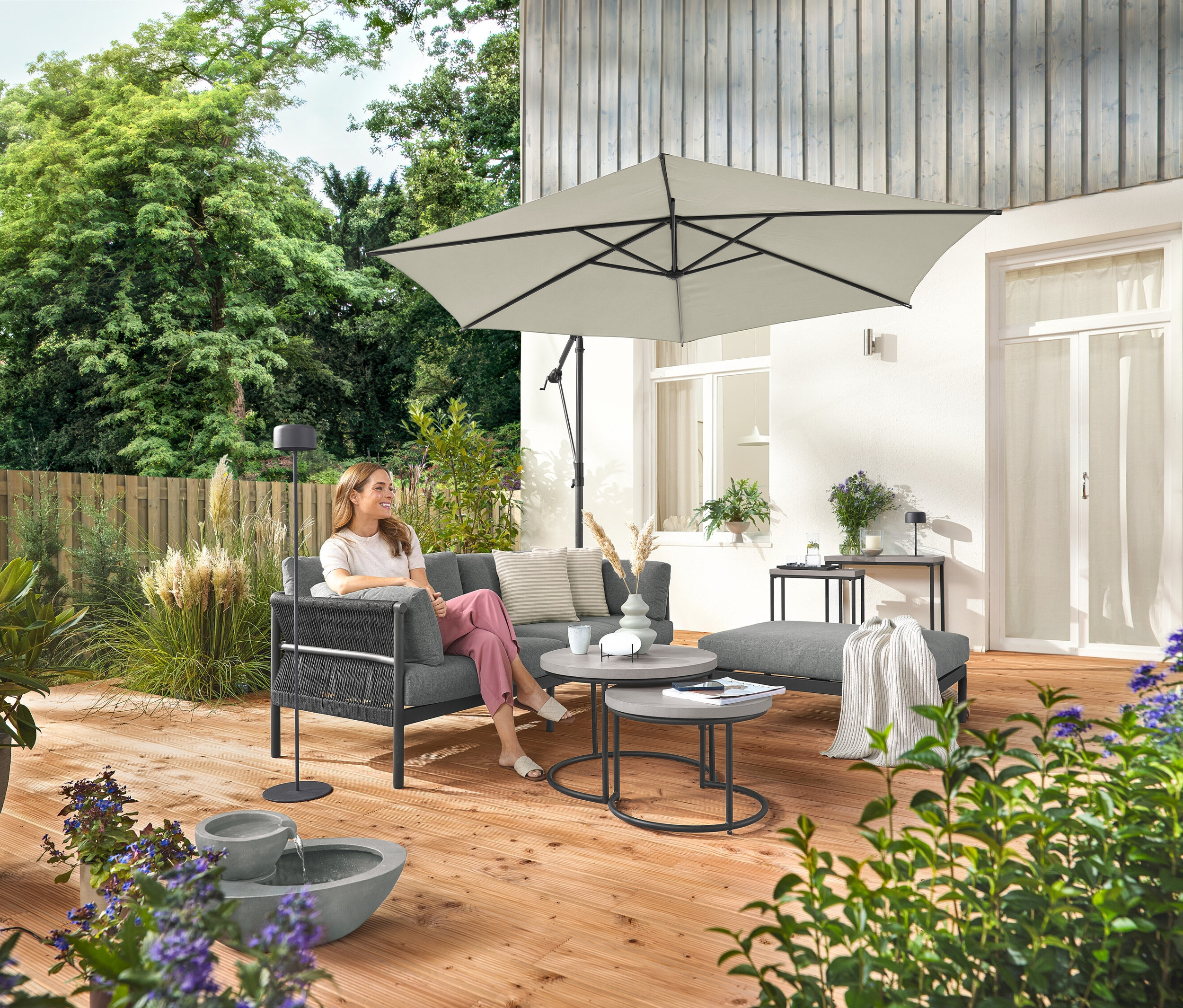 Une femme est assise sur un canapé de jardin confortable avec repose-pieds séparé. Une lampe solaire sur pied grise se trouve à côté d'elle. Au premier plan, une fontaine de jardin solaire.