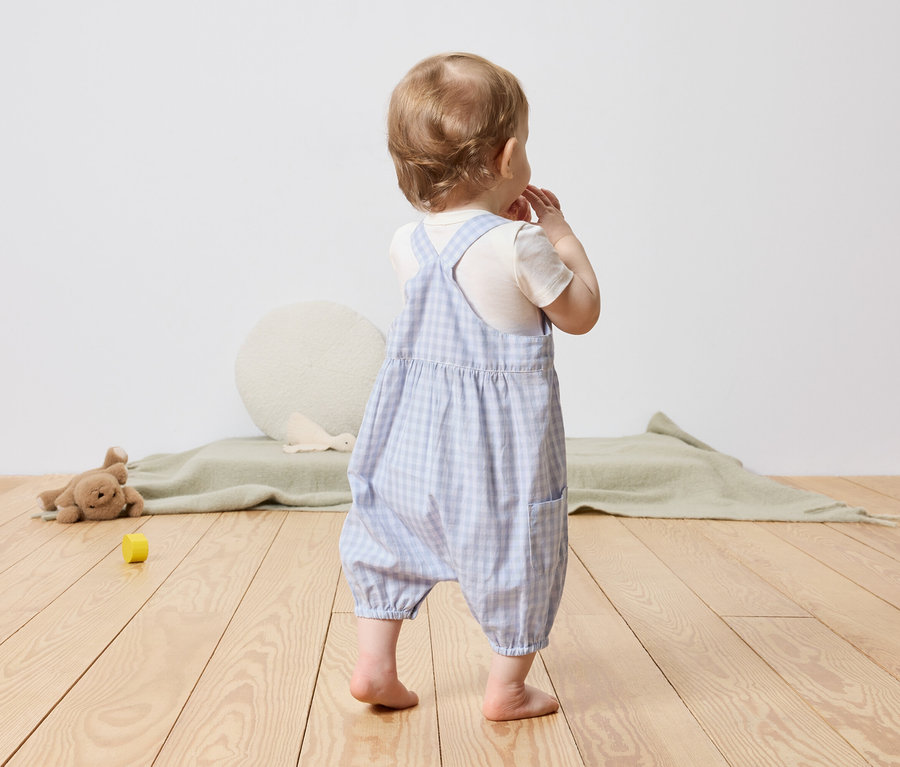 Un jeune enfant se tient dos à la caméra, portant une salopette à carreaux bleus et blancs.