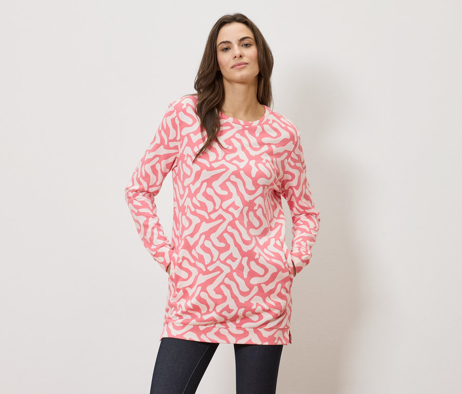 Une femme, les mains dans les poches, porte un haut à manches longues à motifs roses et un jean foncé.