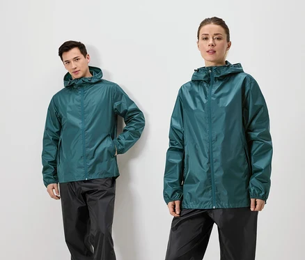 Un homme est appuyé contre un mur et une femme se tient à côté de lui, tous deux portant des vestes de pluie vertes et des pantalons de pluie noirs.