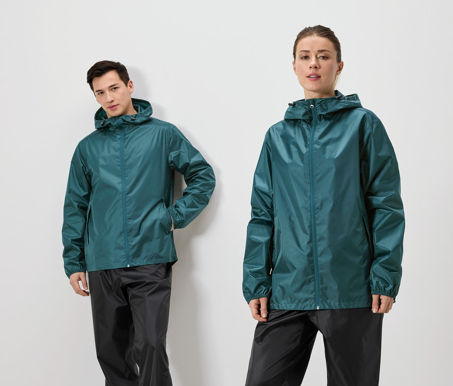 Un homme est appuyé contre un mur et une femme se tient à côté de lui, tous deux portant des vestes de pluie vertes et des pantalons de pluie noirs.