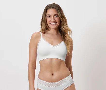 Une femme souriante aux longs cheveux bruns ondulés, portant un soutien-gorge blanc et des sous-vêtements assortis avec des finitions en dentelle.