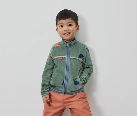 Un garçon souriant aux cheveux bruns porte une veste polaire verte à motifs de dinosaures et un pantalon orange.