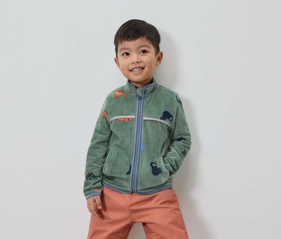Un garçon souriant aux cheveux bruns porte une veste polaire verte à motifs de dinosaures et un pantalon orange.