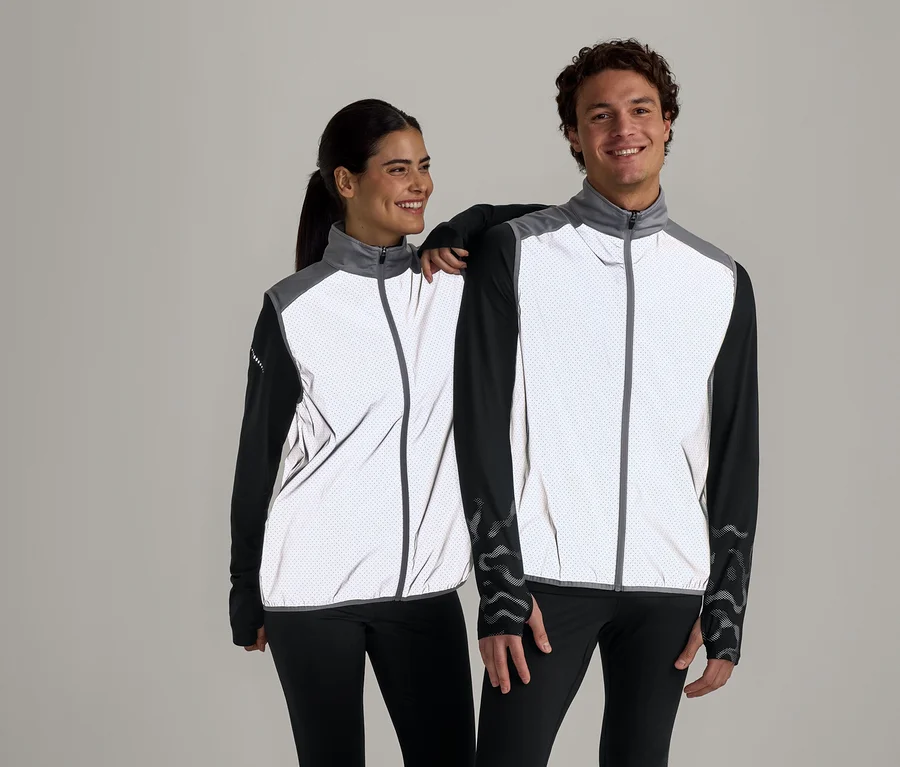Une femme et un homme souriants posent devant un fond gris, vêtus de vestes réfléchissantes à pois blancs avec des manches et des pantalons noirs.
