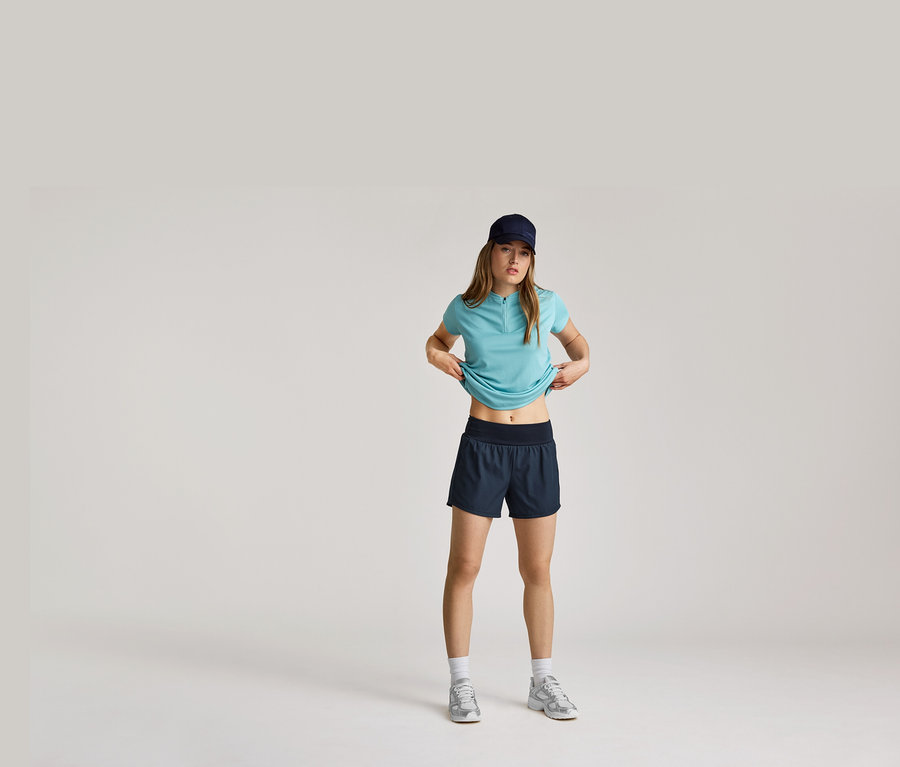 Une femme portant une casquette bleu marine, un t-shirt bleu clair et un short, remontant son t-shirt, avec des chaussettes blanches et des baskets argentées.