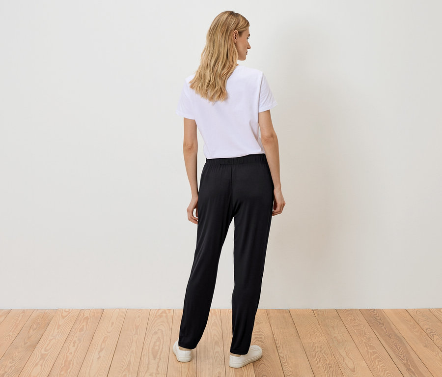 Vue de dos d'une femme aux cheveux blonds ondulés portant un t-shirt blanc, un pantalon noir et des baskets blanches.