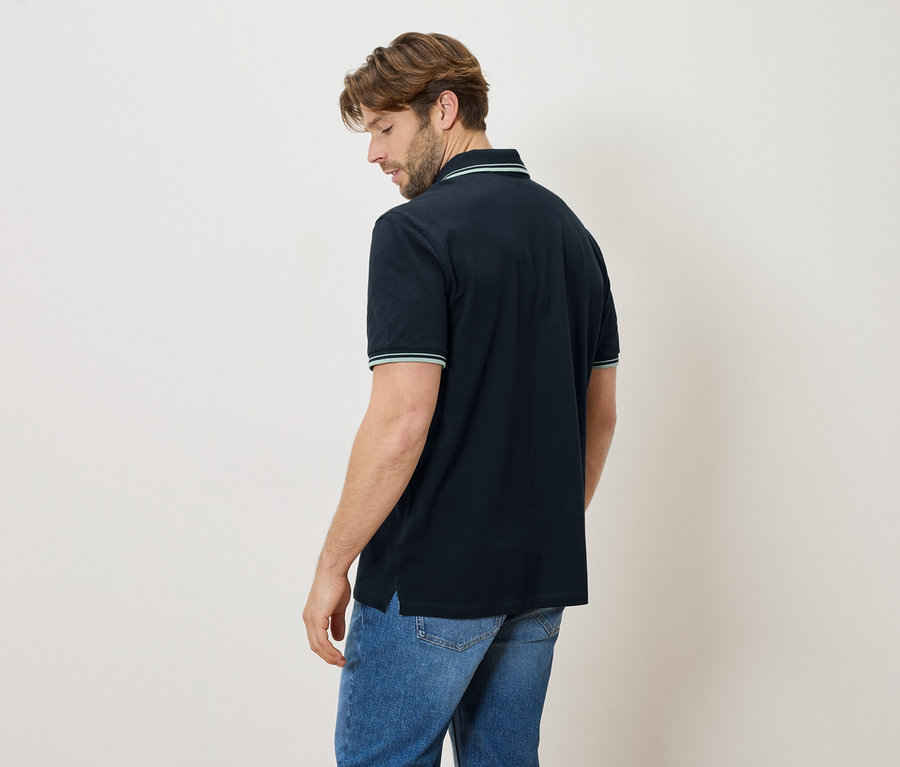 Homme vu de dos portant un polo bleu marine et un jean. Le col et les manches ont des rayures vertes.