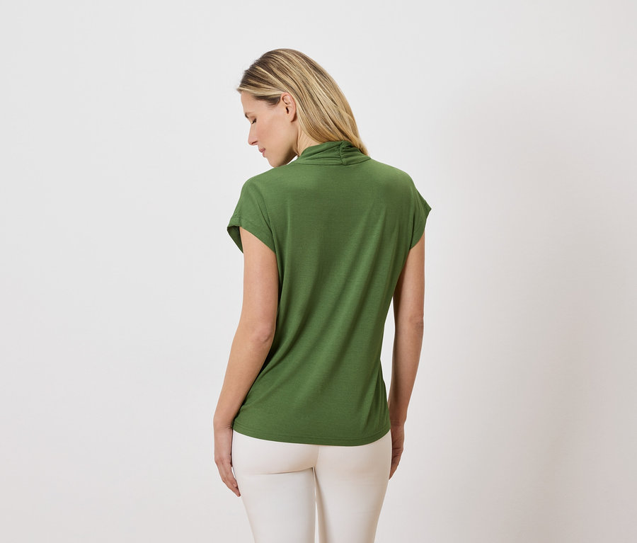 Une femme blonde, vue de dos, porte un t-shirt vert et un pantalon clair sur un fond blanc.