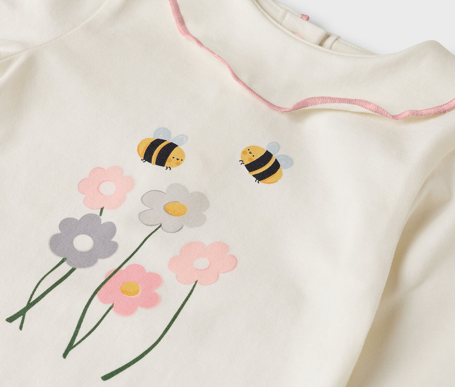 Gros plan sur un haut couleur crème avec un col à volants roses, orné de motifs de fleurs et d'abeilles.