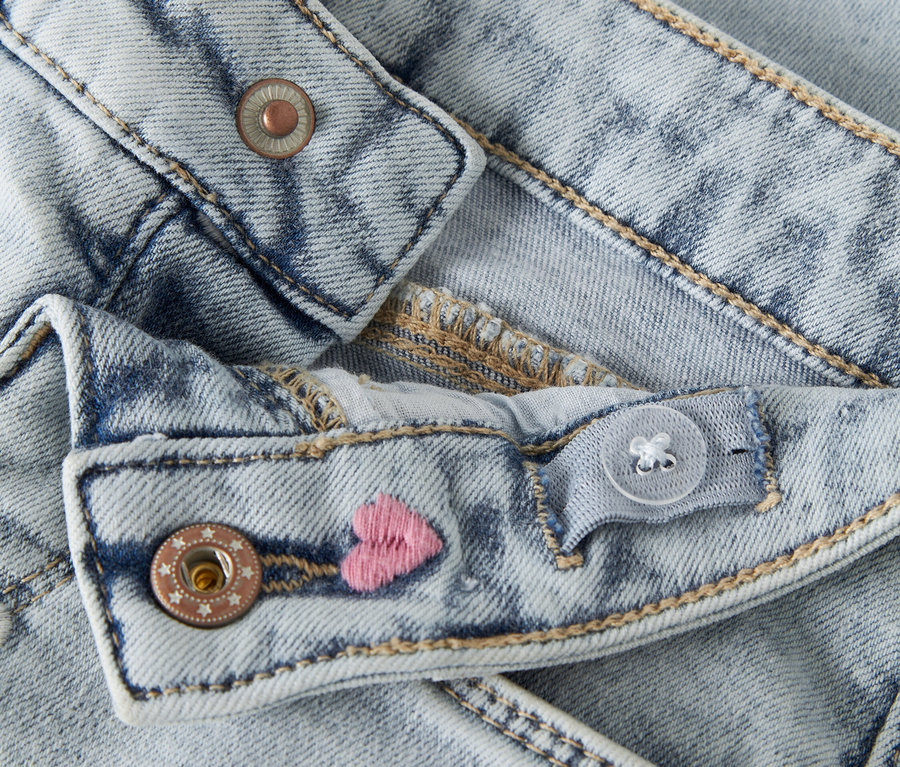 Gros plan sur un jean bleu clair avec une taille ajustable, une fermeture boutonnée et des cœurs roses brodés.