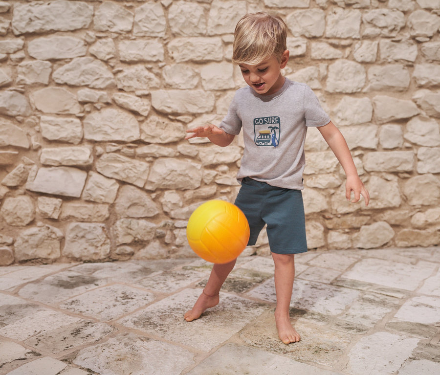 Un garçon joue pieds nus avec un ballon jaune. Il porte un pyjama court pour enfant SCHIESSER avec un t-shirt gris et un short bleu.