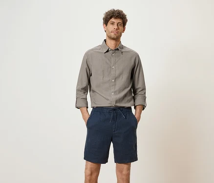 Un homme portant une chemise grise et un short bleu se tient devant un fond blanc.