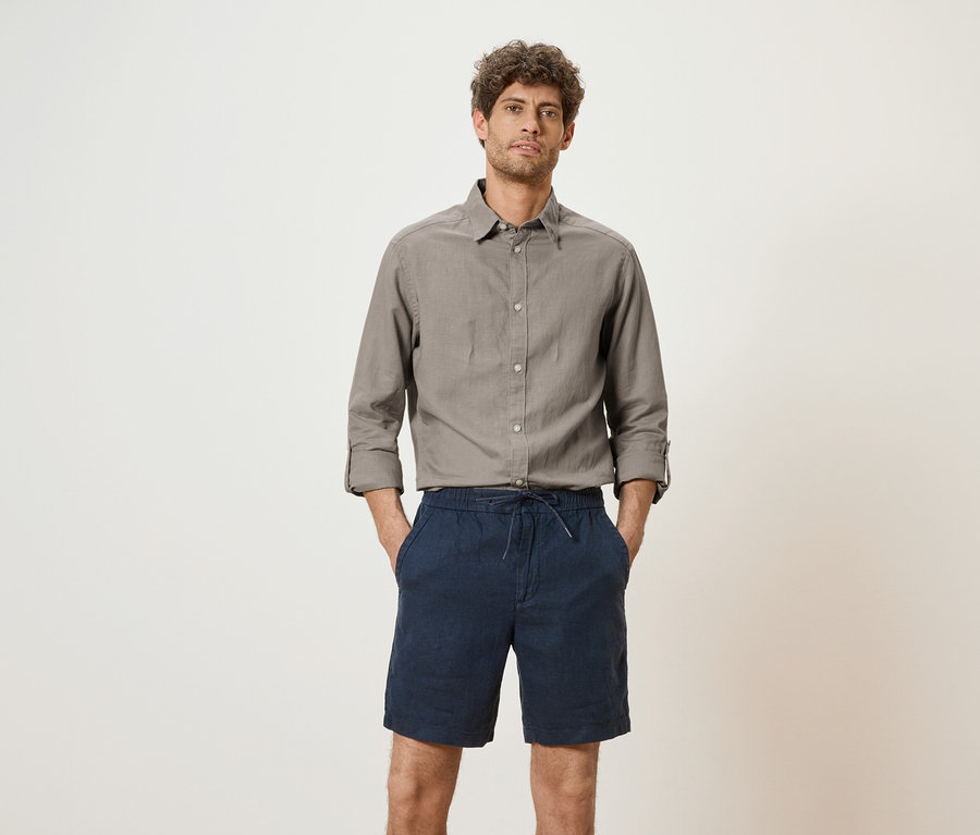 Un homme portant une chemise grise et un short bleu se tient devant un fond blanc.