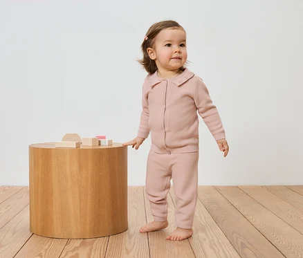 Un jeune enfant portant un cardigan et un pantalon en tricot rose se tient à côté d'une table en bois avec des jouets en bois sur un plancher en bois.