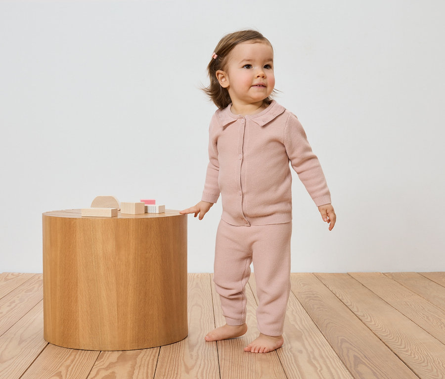 Un jeune enfant portant un cardigan et un pantalon en tricot rose se tient à côté d'une table en bois avec des jouets en bois sur un plancher en bois.