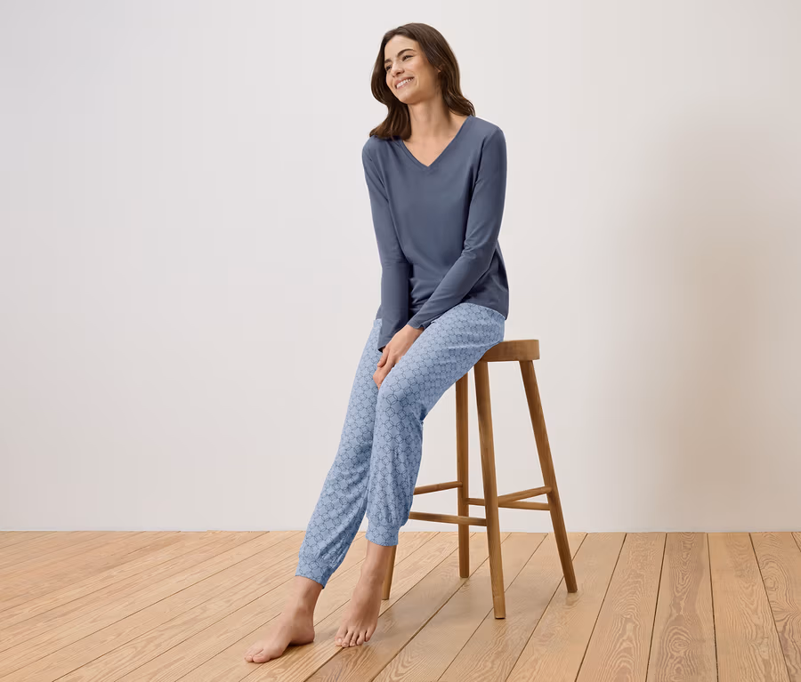 Une femme est assise sur un tabouret et porte un pyjama bleu composé d'un haut à col en V et d'un pantalon à motifs.