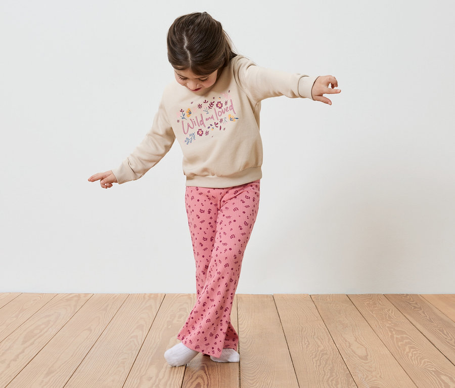 Une fille en équilibre, les bras tendus, sur un plancher en bois. Elle porte un pull beige avec l'inscription "Wild and loved" et un pantalon rose.