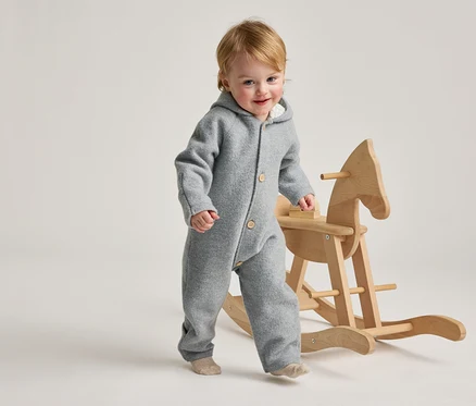 Un bambin souriant aux cheveux blonds, vêtu d'une combinaison grise à capuche et de chaussettes claires, marche à côté d'un cheval à bascule en bois.