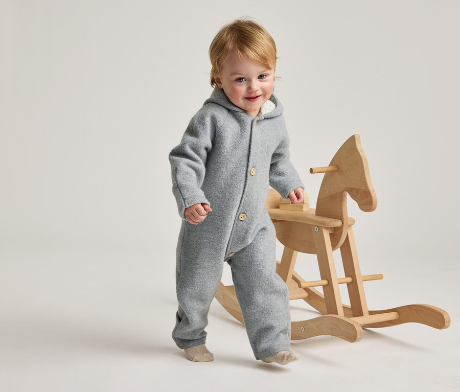 Un bambin souriant aux cheveux blonds, vêtu d'une combinaison grise à capuche et de chaussettes claires, marche à côté d'un cheval à bascule en bois.