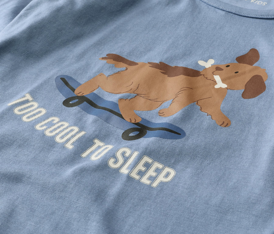 T-shirt bleu clair avec un chien marron allongé sur un skateboard bleu, tenant un os dans sa gueule. En dessous, le texte blanc dit "TOO COOL TO SLEEP".
