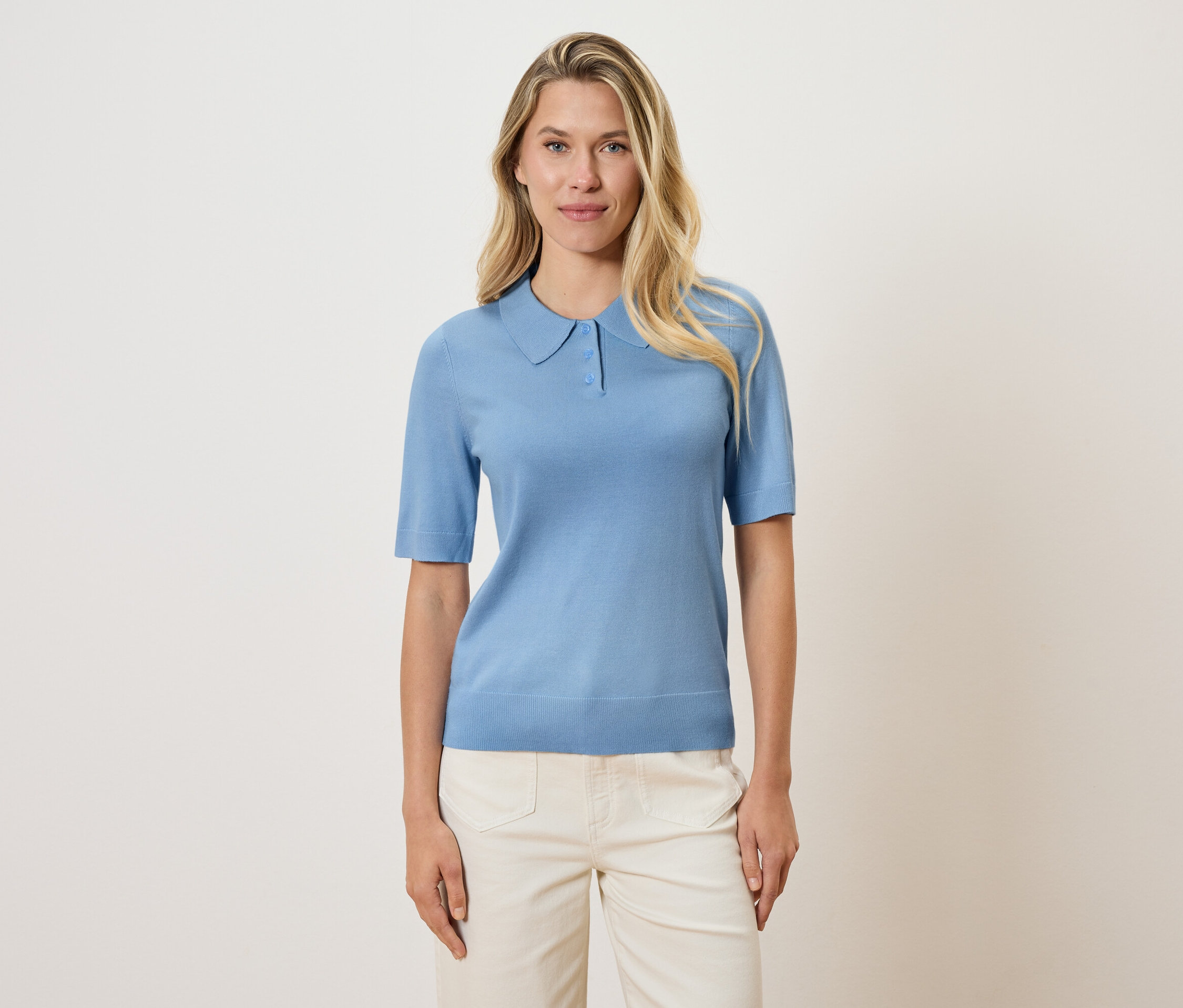Une femme blonde porte un polo bleu clair et un pantalon crème sur un fond crème.