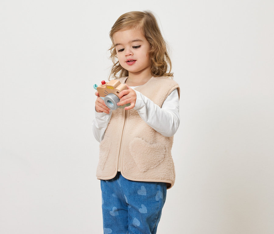 Une petite fille aux cheveux bouclés tient un appareil photo jouet sur un fond clair.