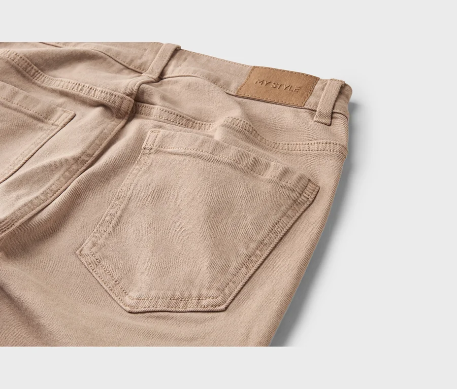 Gros plan du dos d'un pantalon beige, montrant une poche arrière et une étiquette marron « MY STYLE » sur la ceinture.
