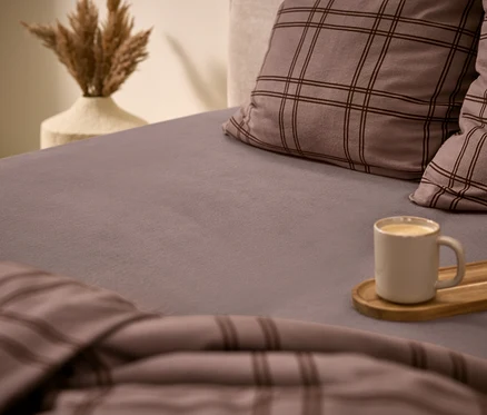 Un lit douillet avec un drap-housse gris, deux oreillers à carreaux gris et une couette assortie. Une tasse blanche avec une boisson est posée sur un plateau en bois sur le lit. À l'arrière-plan, un vase contient de l'herbe de la pampa séchée.
