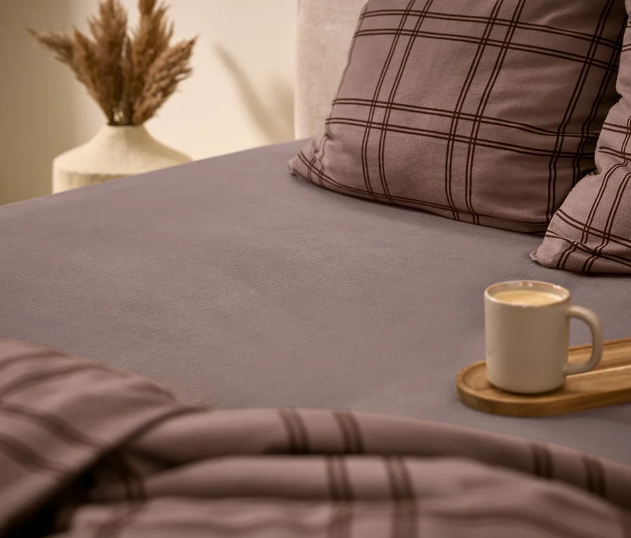 Un lit douillet avec un drap-housse gris, deux oreillers à carreaux gris et une couette assortie. Une tasse blanche avec une boisson est posée sur un plateau en bois sur le lit. À l'arrière-plan, un vase contient de l'herbe de la pampa séchée.