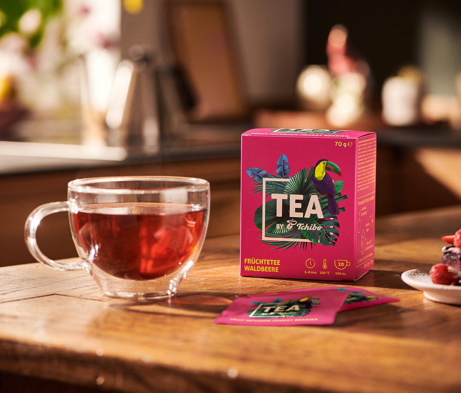 Sur une table en bois se trouvent une tasse en verre de tisane, un paquet de TEA by Tchibo infusion aux fruits des bois et une assiette de dessert.