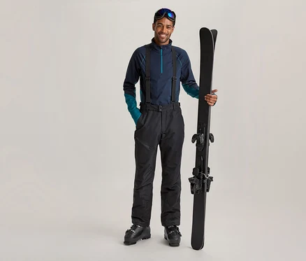 Homme souriant en tenue de ski bleu foncé avec des bretelles et des lunettes de ski sur le front, tenant des skis noirs à la verticale.