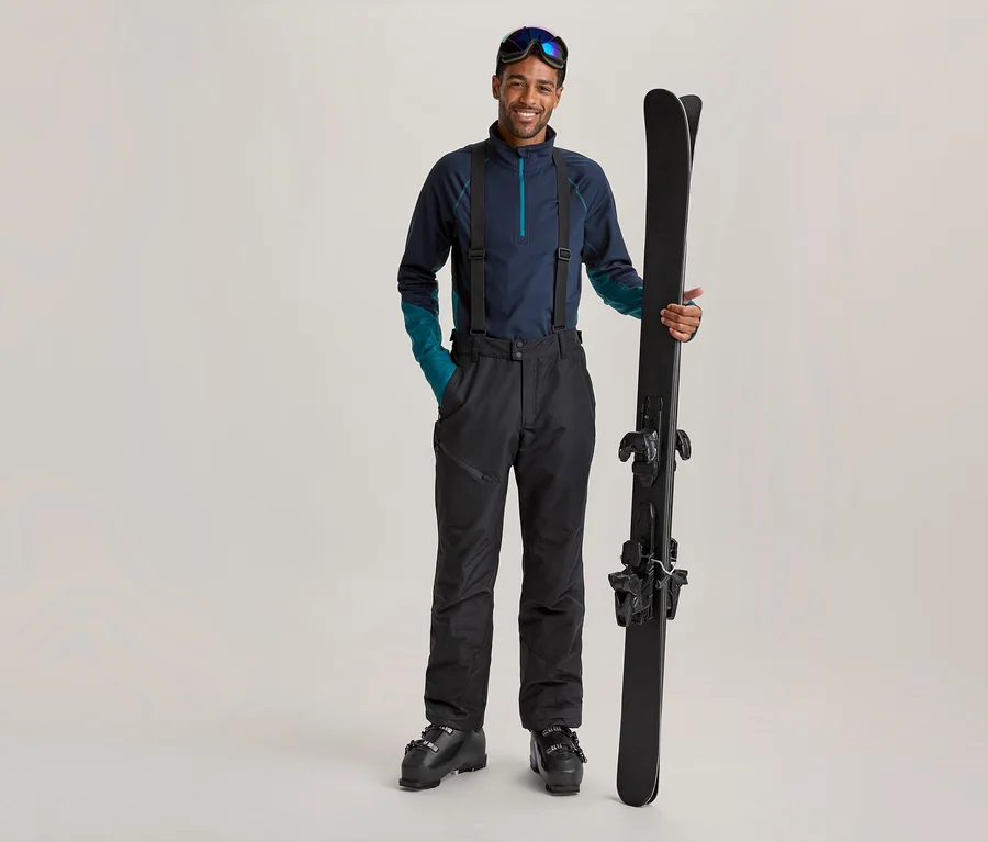 Homme souriant en tenue de ski bleu foncé avec des bretelles et des lunettes de ski sur le front, tenant des skis noirs à la verticale.