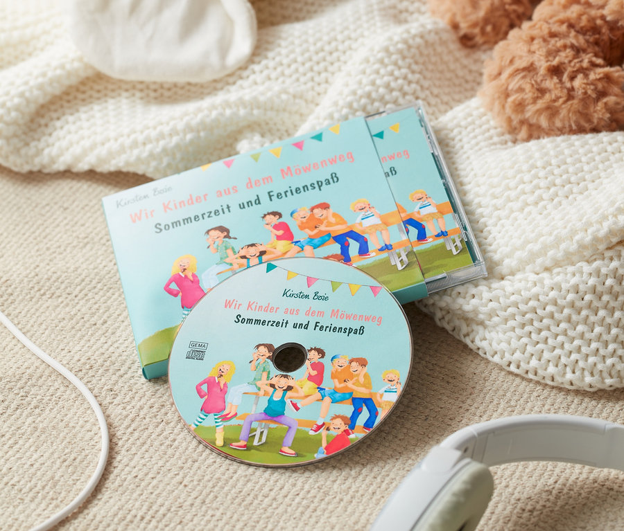 Un livre audio « Wir Kinder aus dem Möwenweg – Sommerzeit und Ferienspaß » et des écouteurs sont posés sur une couverture à côté d'une peluche.