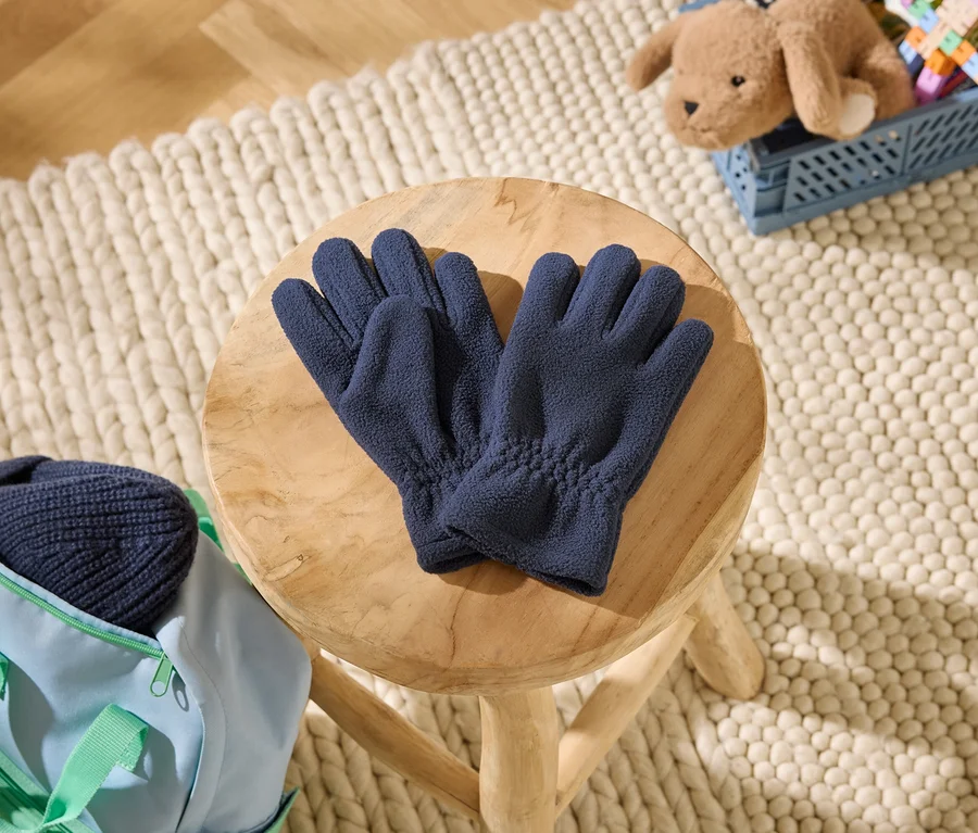 Des gants polaires bleu foncé sont posés sur un tabouret en bois. À côté, un sac bleu clair avec un bonnet bleu foncé. À l'arrière-plan, un ours en peluche dans une caisse bleue sur un tapis.