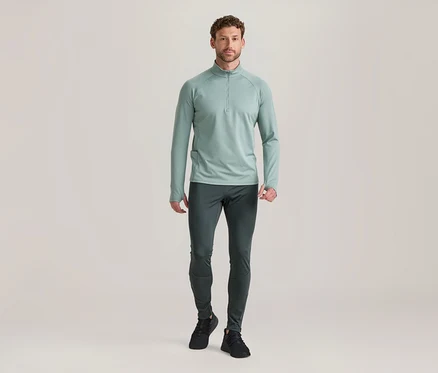 Homme debout, portant un haut à manches longues vert clair avec demi-zip, un pantalon de sport gris foncé et des baskets noires.