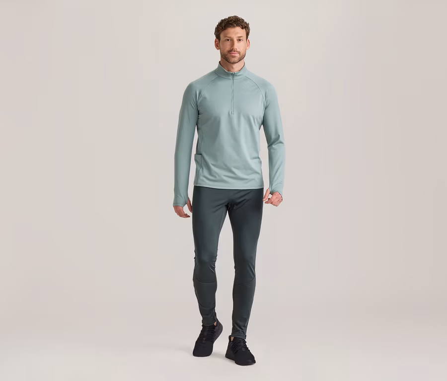 Homme debout, portant un haut à manches longues vert clair avec demi-zip, un pantalon de sport gris foncé et des baskets noires.