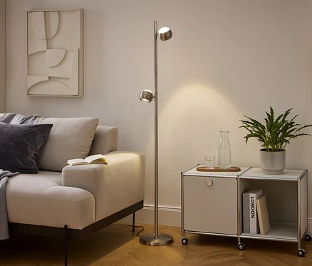 Un salon moderne avec un canapé beige, un lampadaire en métal brossé et un meuble modulaire. Le canapé est orné d'un plaid et d'un coussin gris, avec un livre ouvert sur son accoudoir. La lampe éclaire la zone à côté d'un meuble contenant une plante, une carafe d'eau et des livres.