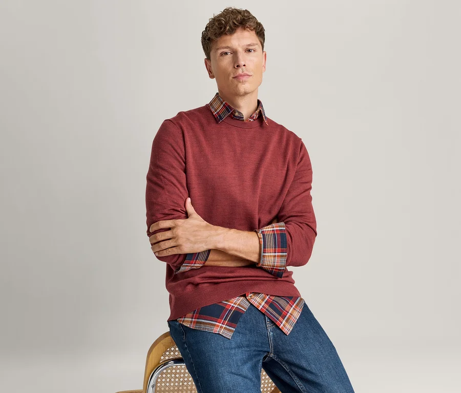 Homme assis les bras croisés, portant un pull rouge à manches longues sur une chemise à carreaux rouge, bleue et orange, et un jean bleu.