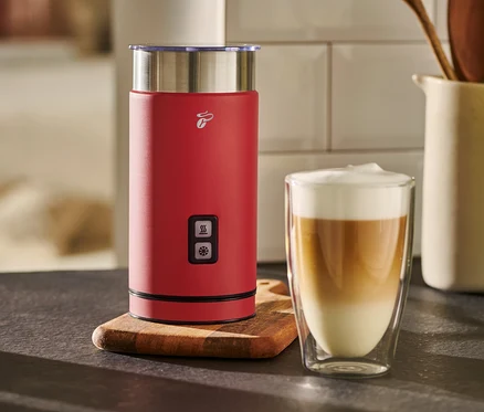 Un mousseur à lait électrique rouge se trouve à côté d'un verre de café au lait.
