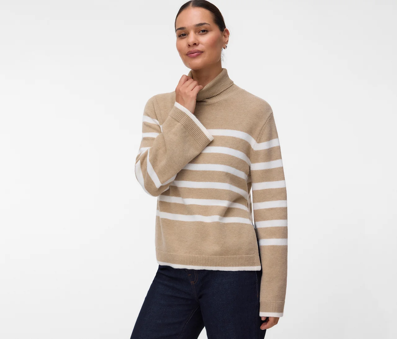 Femme portant un pull à col roulé rayé beige et blanc et un jean foncé, regardant la caméra avec une main près du cou.