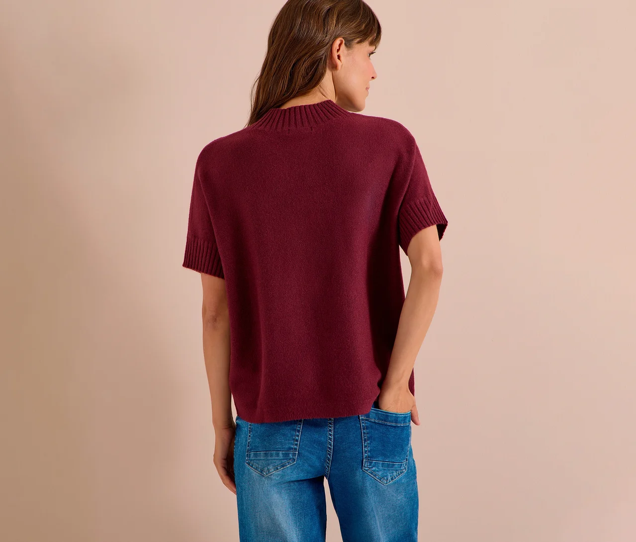 Femme de dos, portant un pull en maille CECIL rouge bordeaux à manches courtes et un jean bleu. Sa main droite est dans la poche de son jean.