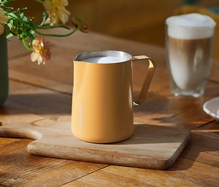 Un pichet à lait jaune rempli de lait moussé est posé sur une planche à découper en bois sur une table en bois. À l'arrière-plan, on voit un verre de latte, un vase avec des fleurs claires et une assiette de biscuits.