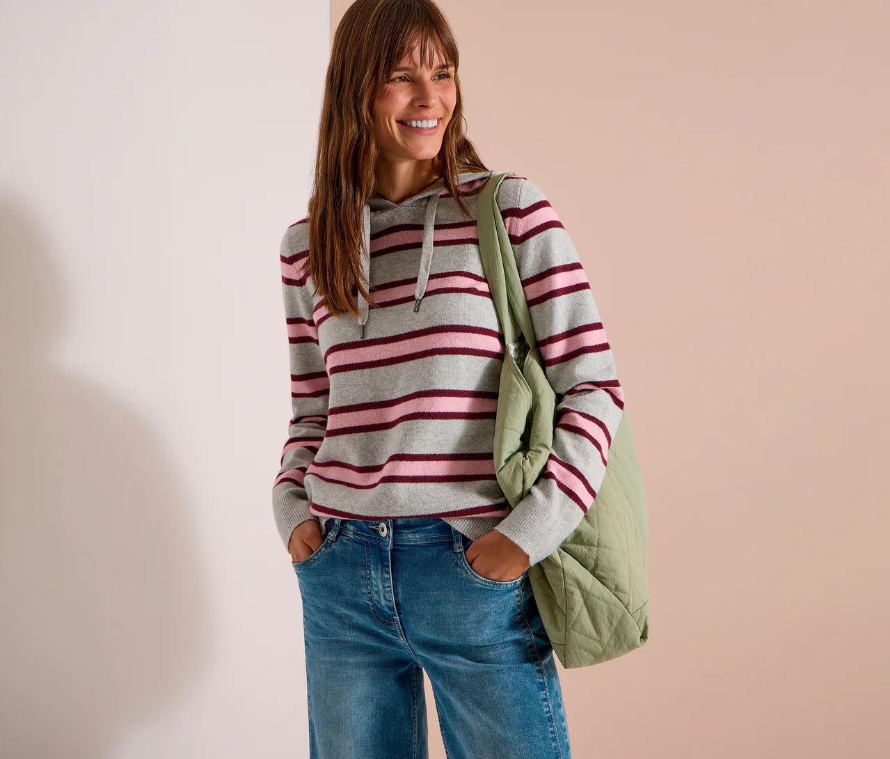 Une femme souriante aux longs cheveux bruns porte un sweatshirt à capuche en maille CECIL gris, rose et bordeaux à rayures, un jean bleu et un sac fourre-tout matelassé vert clair. Ses mains sont dans ses poches.