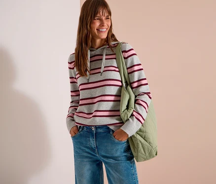 Une femme souriante aux longs cheveux bruns porte un sweatshirt à capuche en maille CECIL gris, rose et bordeaux à rayures, un jean bleu et un sac fourre-tout matelassé vert clair. Ses mains sont dans ses poches.
