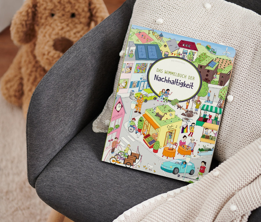 Sur un fauteuil gris reposent un plaid en tricot beige et le livre « Das Wimmelbuch der Nachhaltigkeit ». Un ours en peluche est visible en arrière-plan.