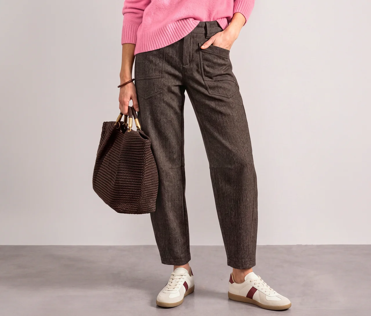 Une personne porte un pull rose, un pantalon en tissu Street One marron foncé et des baskets blanches à rayures rouges, tenant un sac tressé marron et ayant une main dans la poche.