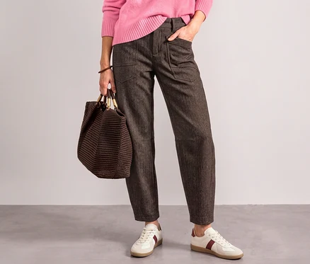 Une personne porte un pull rose, un pantalon en tissu Street One marron foncé et des baskets blanches à rayures rouges, tenant un sac tressé marron et ayant une main dans la poche.